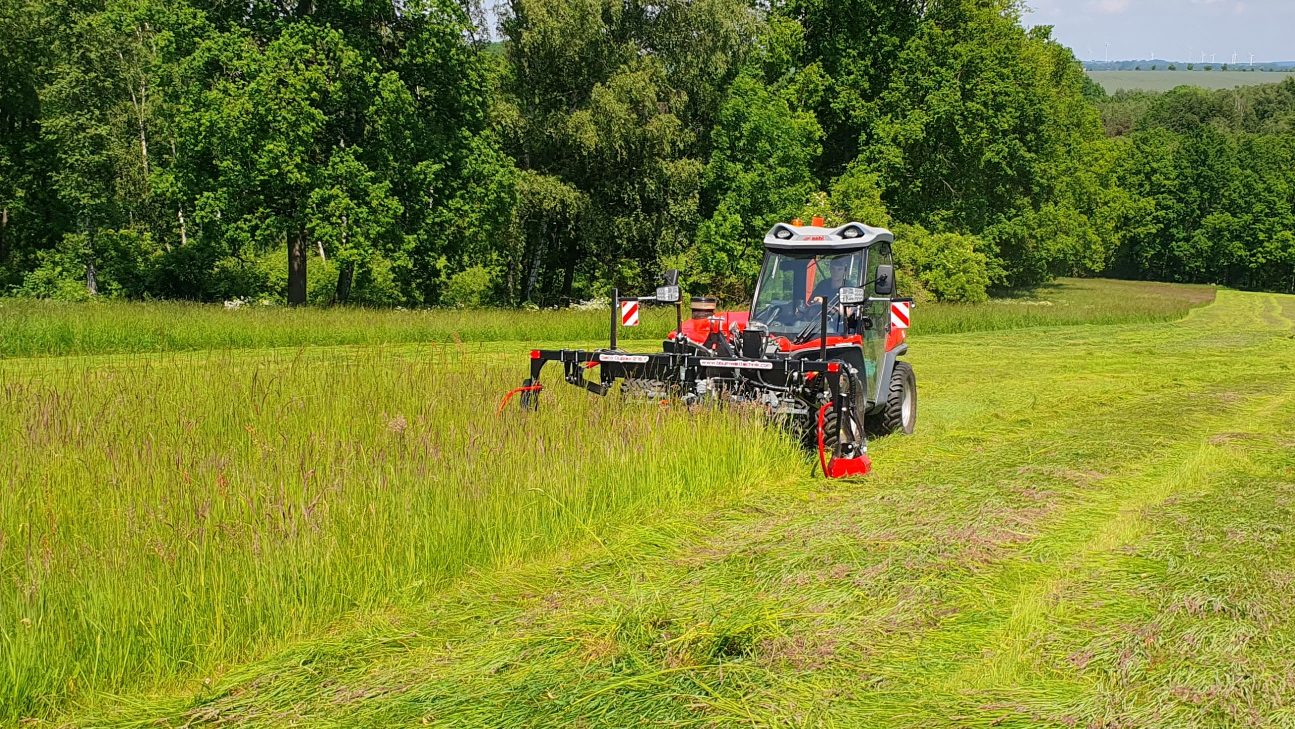 Ein Traktor mäht insektenschonend Gras mit einem Doppelmessermähwerk auf einer Wiese in einer grünen Landschaft.