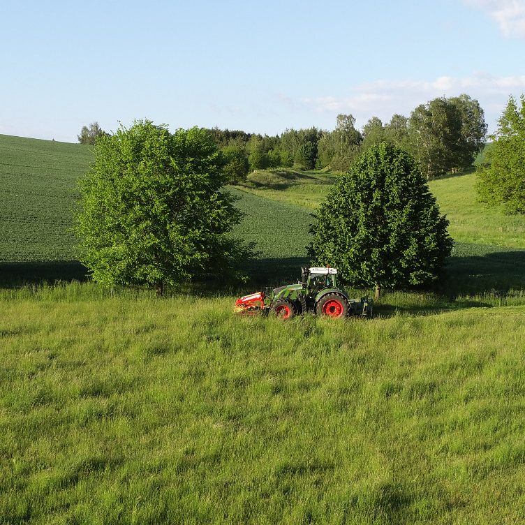 Traktor mäht Wiese in ländlicher Umgebung mit Bäumen und sanften Hügeln.